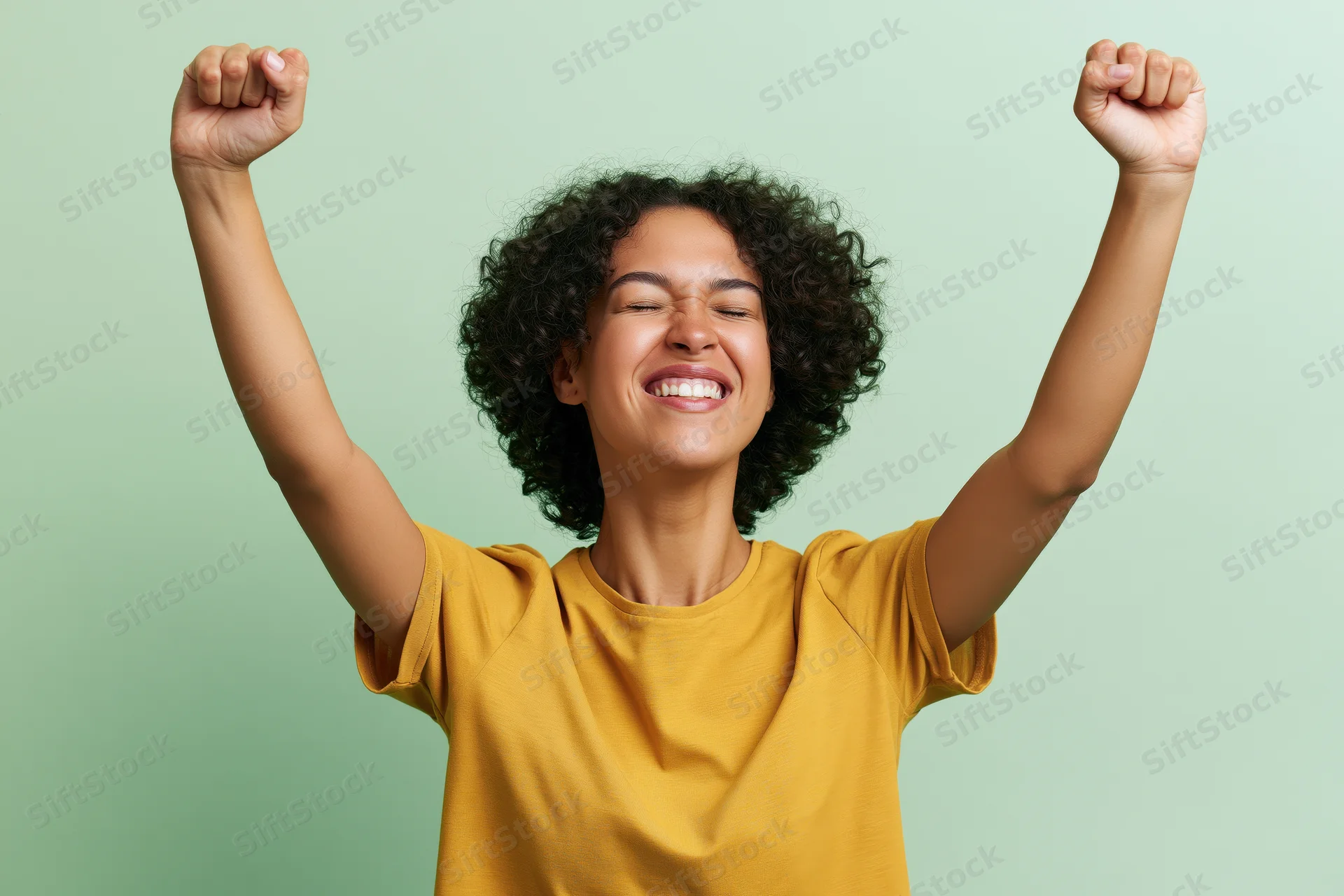 Young brazilian woman laughing with eyes closed and arms raised in triumph against green background