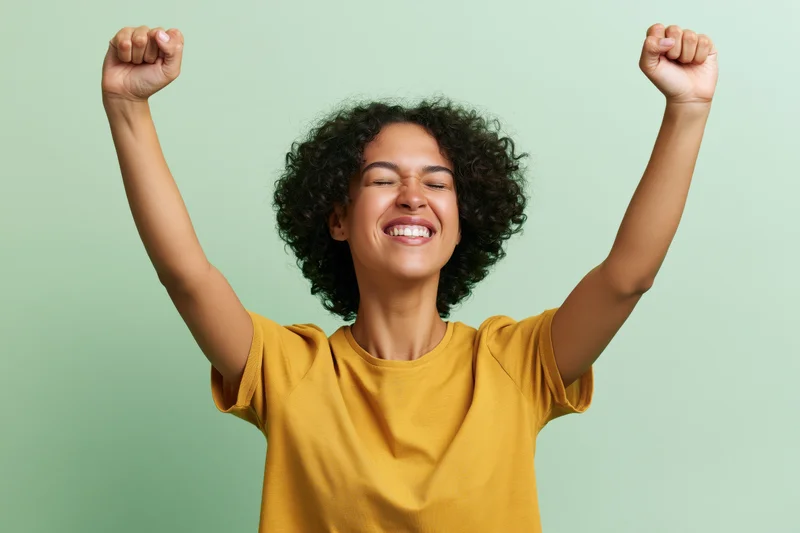 Young brazilian woman laughing with eyes closed and arms raised in triumph against green background