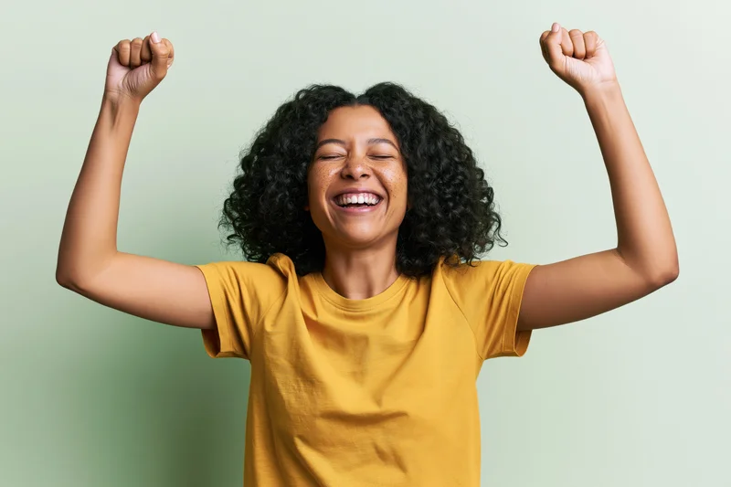 Young woman laughing with eyes closed and fists raised in celebration against a green background