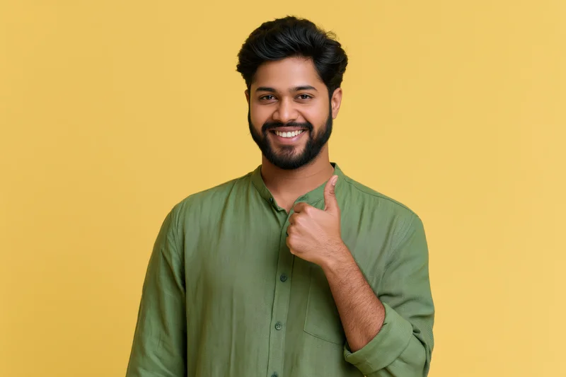 Young indian man smiling and pointing with thumb to the side against yellow background