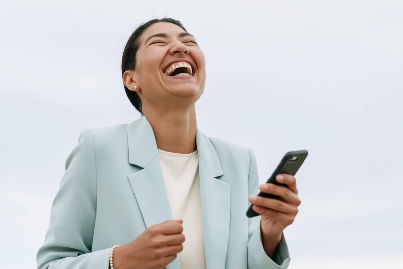 Young latina woman laughing joyfully while holding a smartphone outdoors