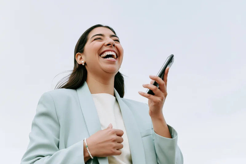 Young latina woman laughing joyfully while holding smartphone outdoors