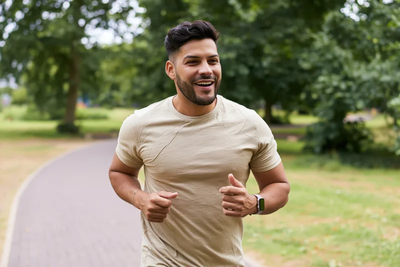 Young latino man jogging on a paved path in a city park smiling with trees and greenery around
