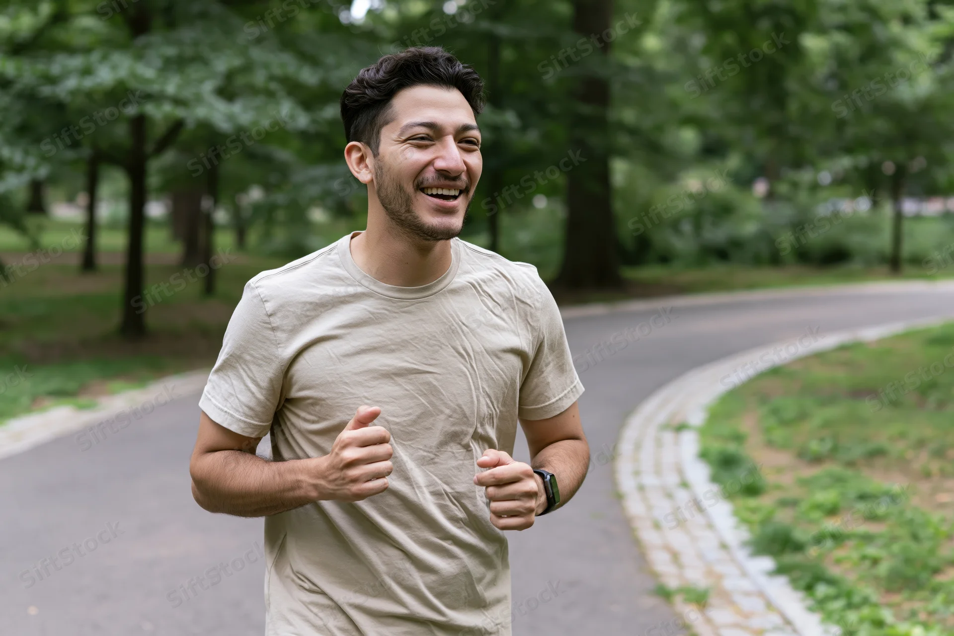Young latino man jogging through a city park laughing and moving along a paved path surrounded by trees