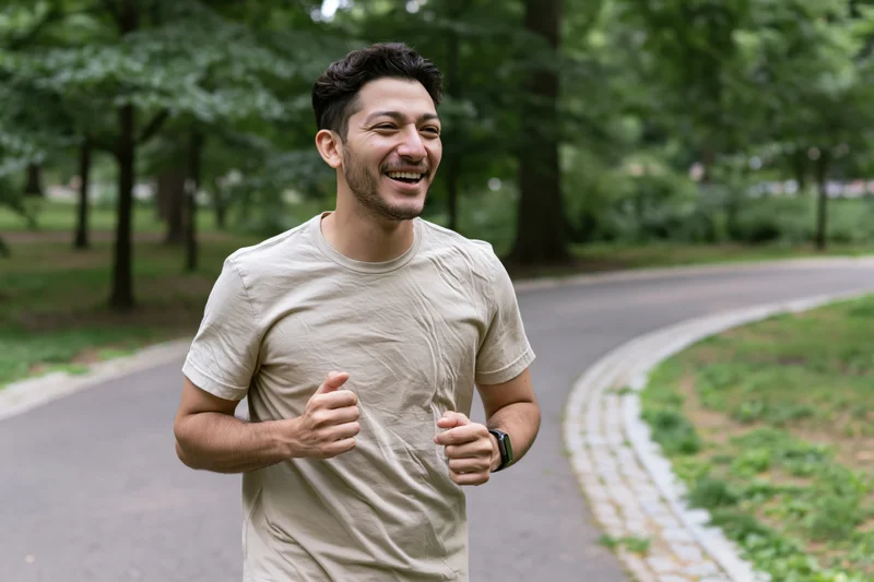 Young latino man jogging through a city park laughing and moving along a paved path surrounded by trees