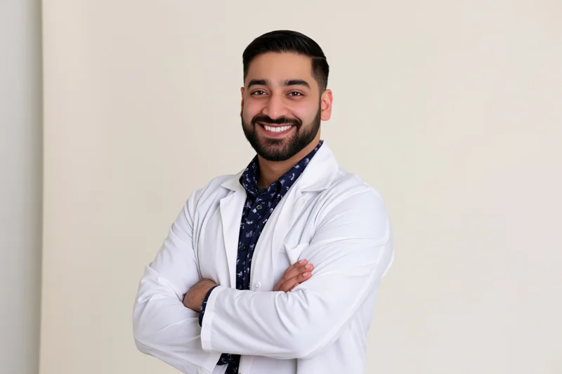 Young southasian male doctor in white lab coat smiling with arms crossed against plain wall