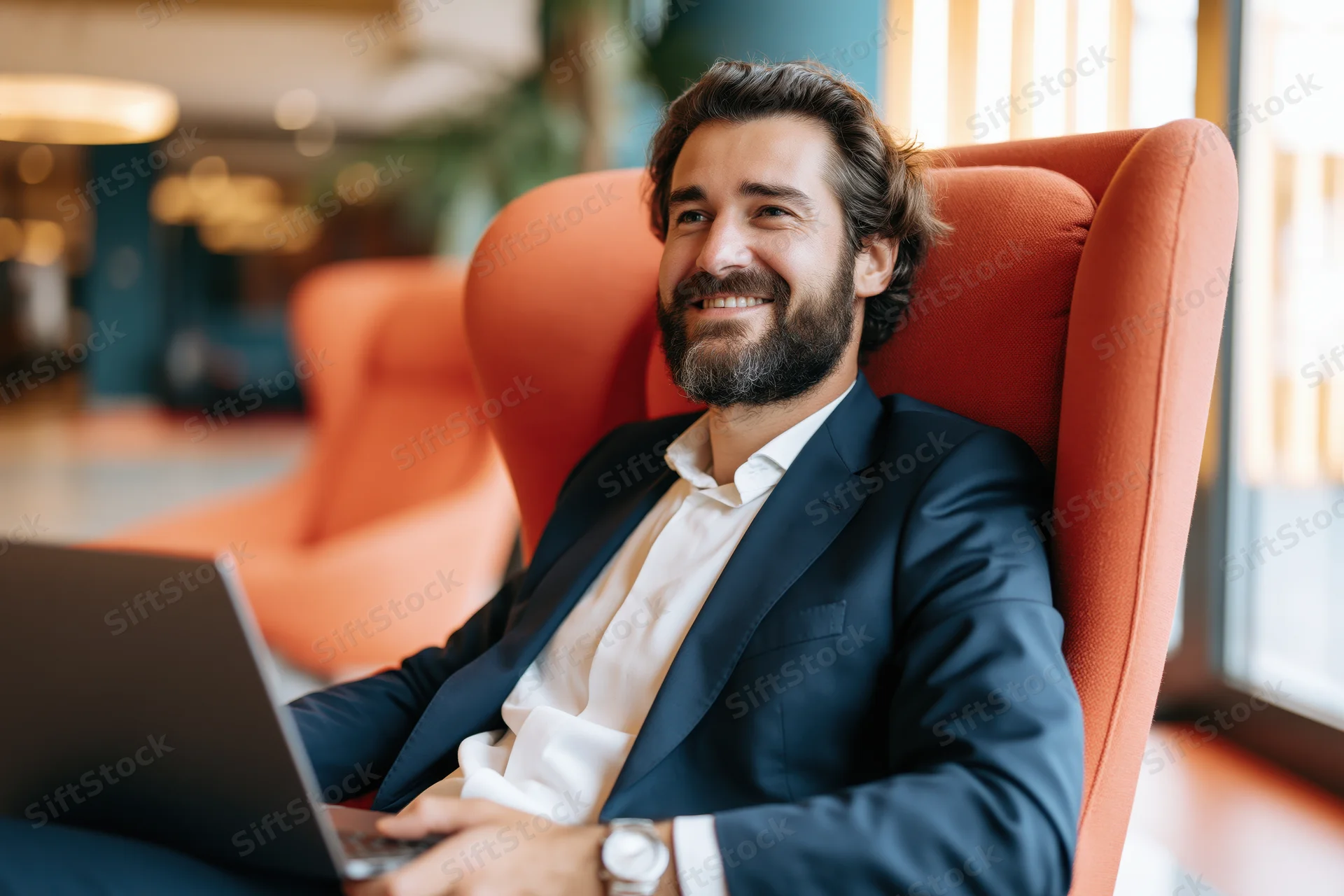 Bearded businessman smiling while using laptop in orange high-back chair in modern office space