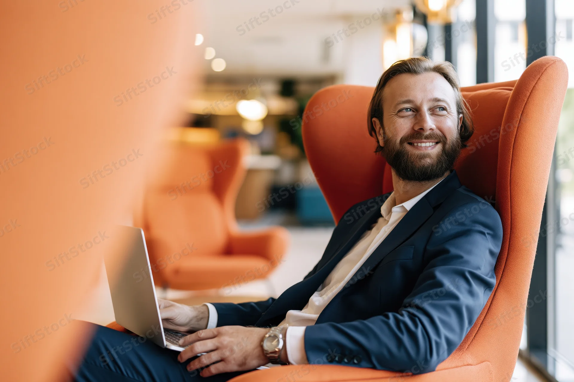 Young bearded businessman smiling while using laptop in orange high-back chair in modern office