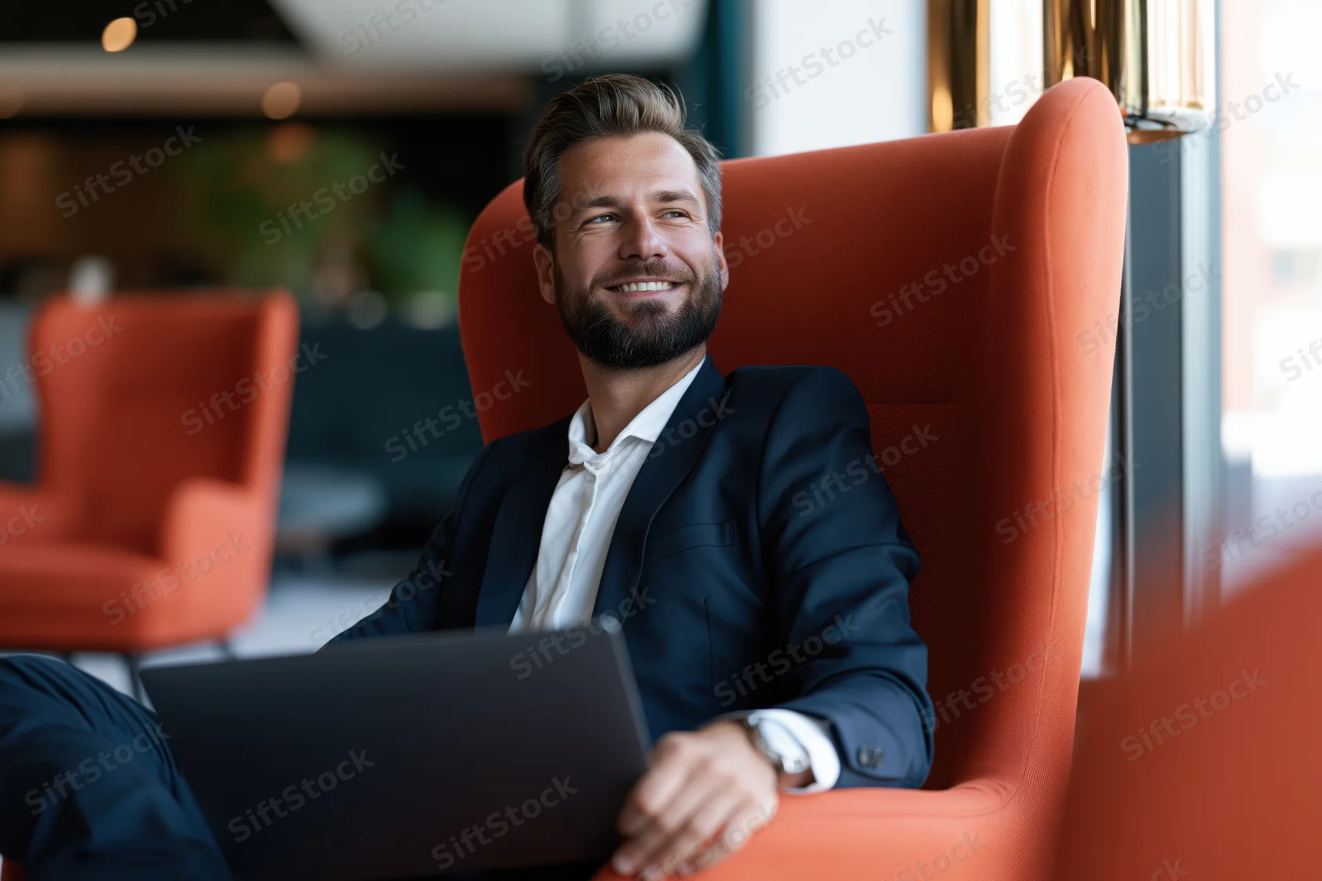 Businessman smiling while sitting in orange armchair with laptop in modern office lounge