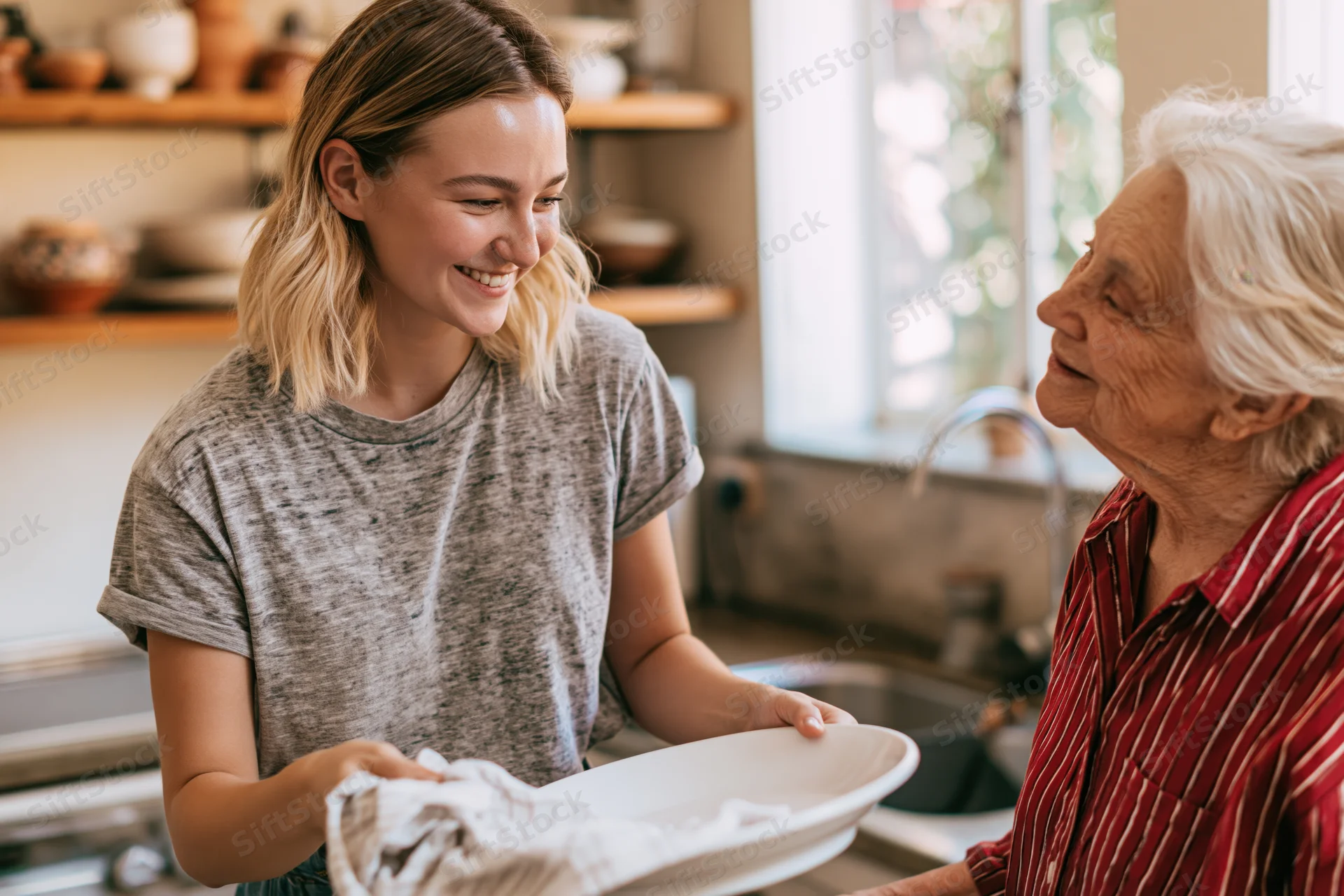 Young woman drying a white plate with a cloth while smiling at an elderly woman in a kitchen