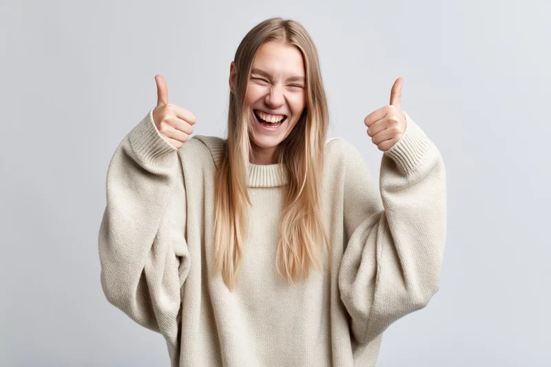 Young blonde woman laughing and giving two thumbs up in a beige sweater against a light gray background