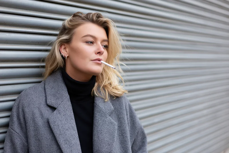Young blonde woman smoking a cigarette while leaning against a metal shutter outdoors