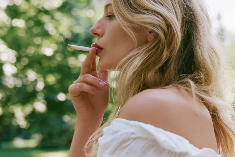 Blonde woman smoking a cigarette outdoors in profile with green foliage background