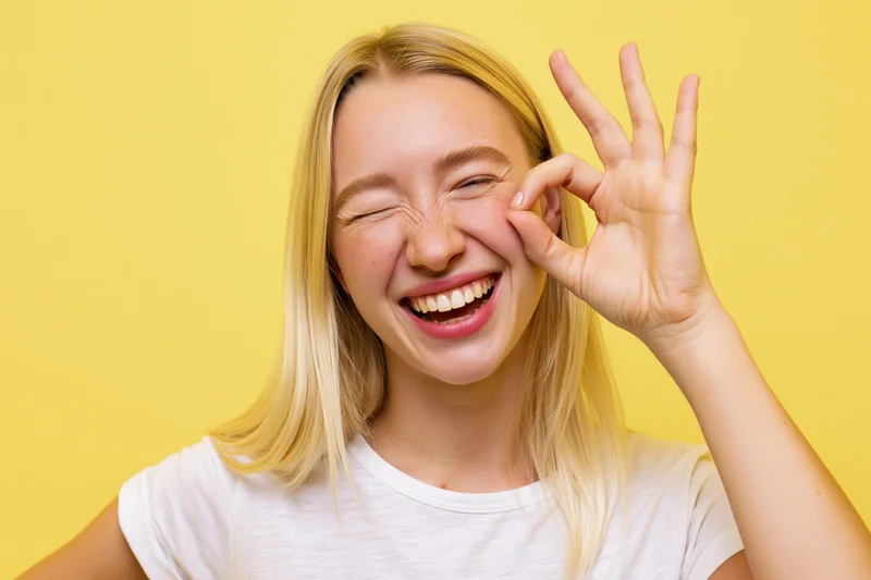 Young blonde woman laughing and winking with ok hand gesture against yellow background
