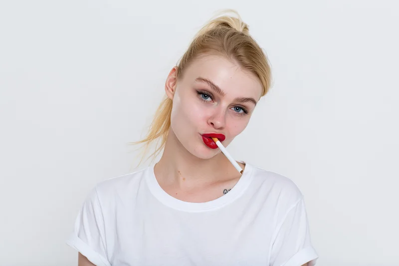Young blonde woman with high ponytail holding a cigarette in her mouth, wearing white t-shirt, in studio setting