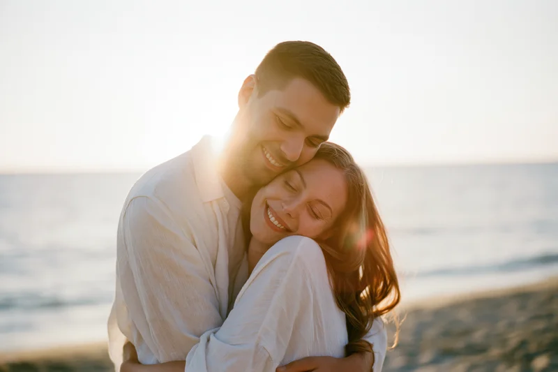 Young couple embracing on a beach at sunset, smiling and hugging closely with ocean in background