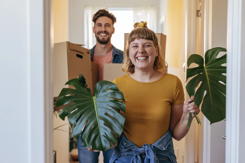Woman laughing while holding monstera leaves in front of man carrying boxes in apartment hallway