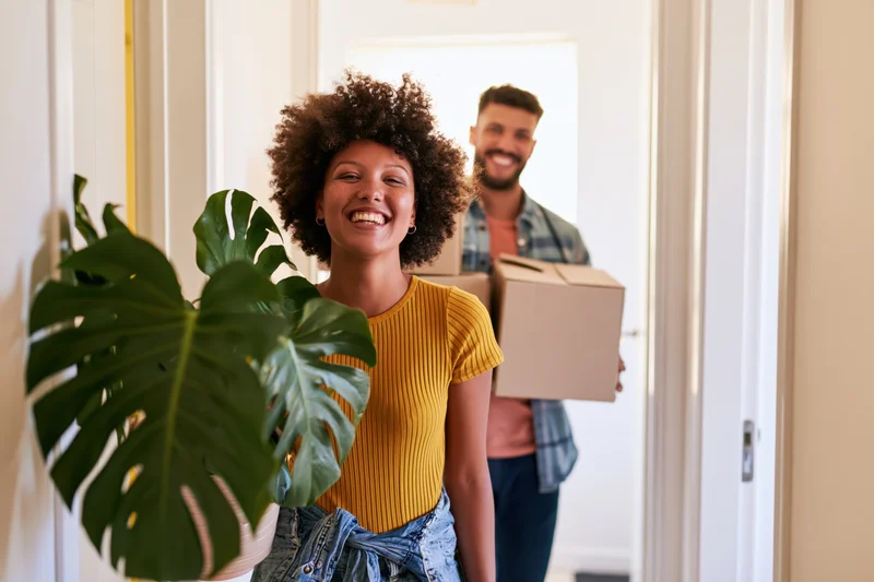 Woman smiling in front of doorway holding plant as man behind carries boxes into new apartment