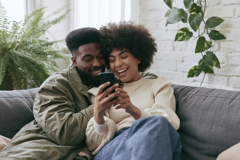 Young couple laughing together while looking at smartphone on grey sofa at home