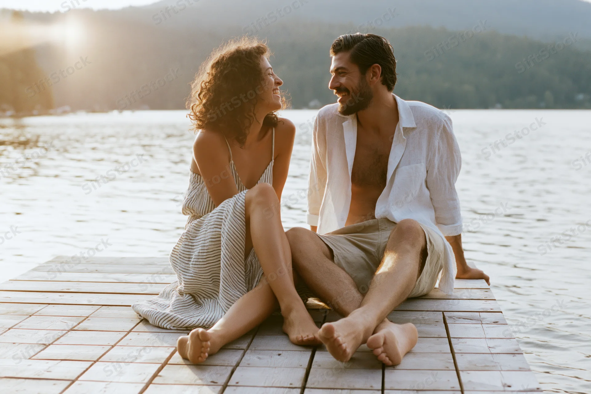 Young couple laughing barefoot on wooden dock by lake at sunset