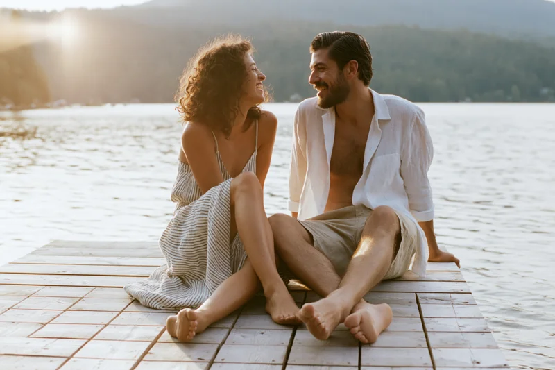 Young couple laughing barefoot on wooden dock by lake at sunset