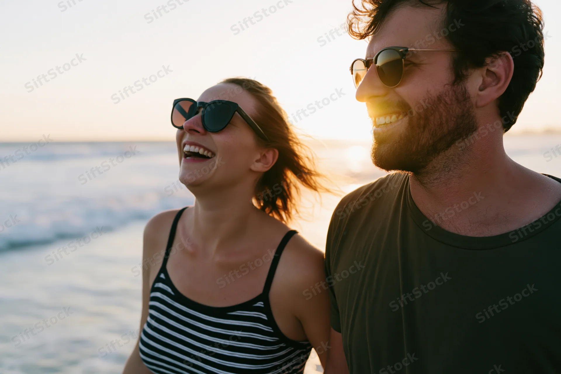 Young couple laughing while walking arm in arm along the beach at sunset