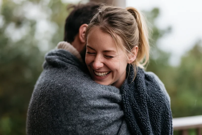 Woman smiling while hugging man wrapped in grey wool blanket outdoors