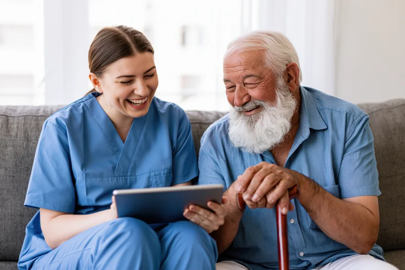 Female caregiver in blue scrubs laughing with elderly man while showing tablet on couch