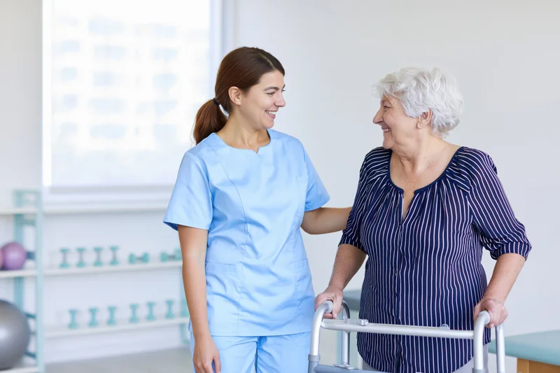 Young female caregiver supporting senior woman with walker in rehabilitation clinic