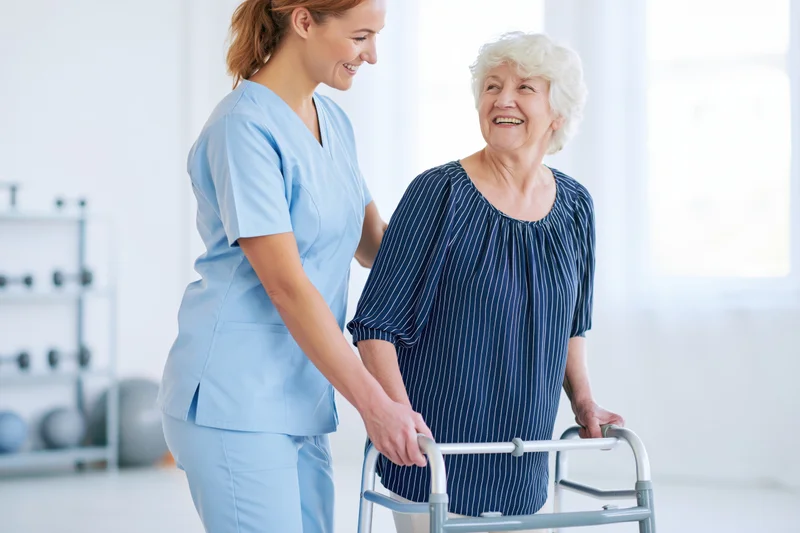 Young female caregiver in light blue scrubs supporting an elderly woman with a walker in a bright clinic