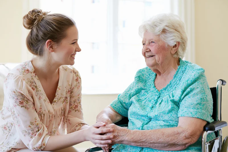 Young female caregiver kneeling beside an elderly woman in a wheelchair, holding her hands and smiling at each other in a sunlit room