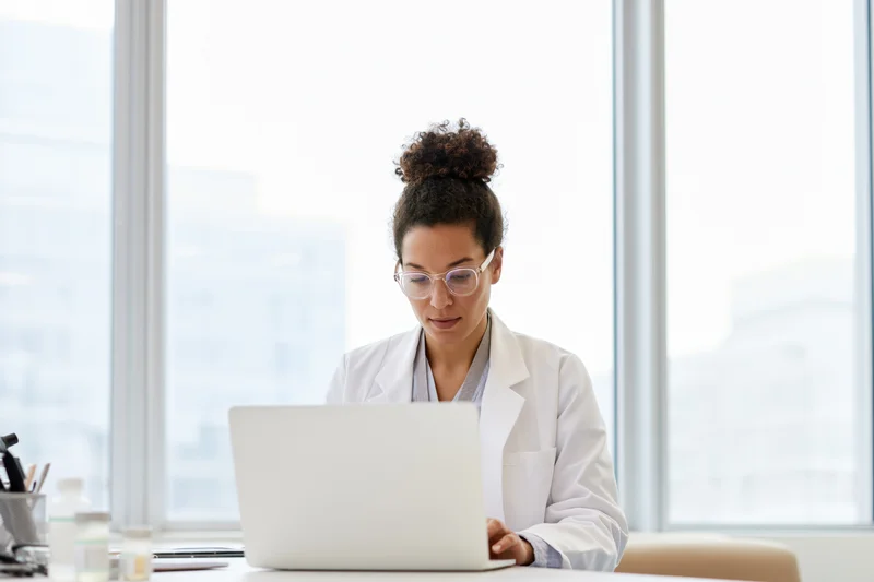 Woman in white lab coat working on laptop at desk in bright office