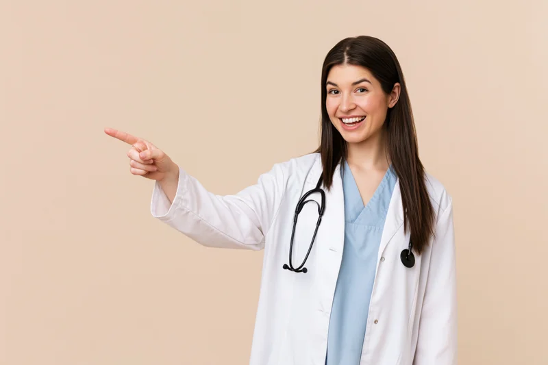 Young female doctor in white coat pointing to the left while smiling, beige background