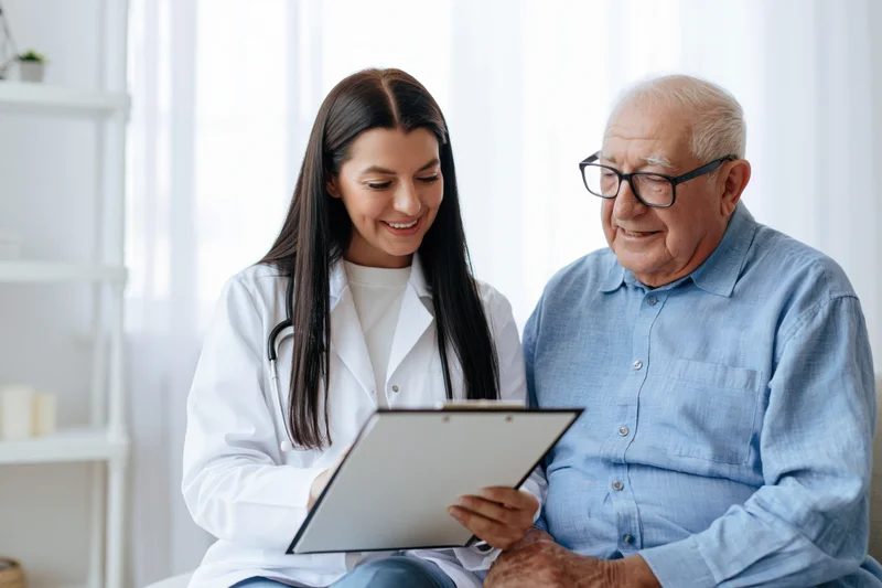Young female doctor consulting with senior patient in clinic, holding clipboard and smiling
