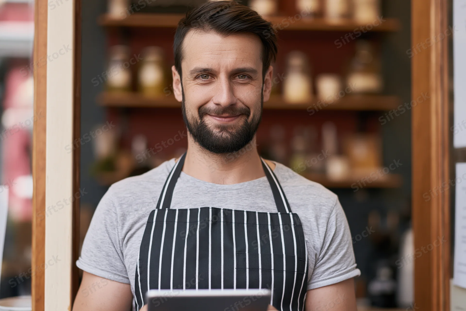 Smiling male cafe owner in striped apron holding tablet at shop entrance