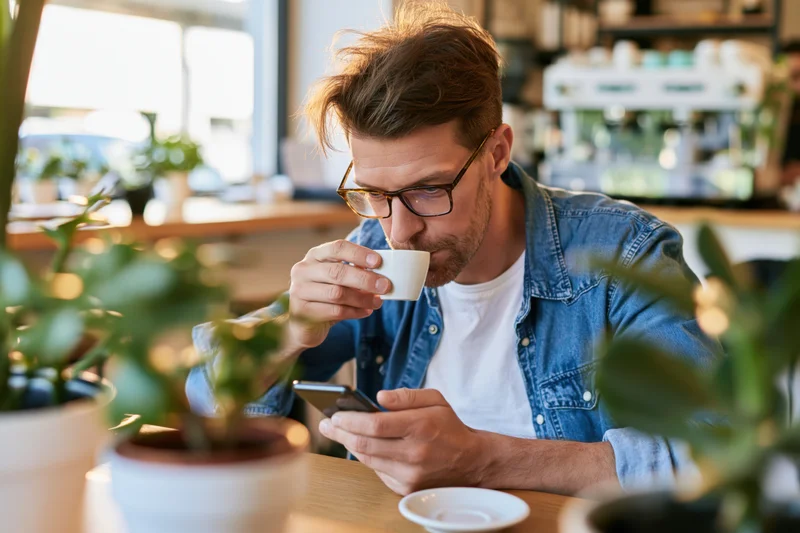 Man sipping espresso from a white cup while holding a smartphone at a cafe table
