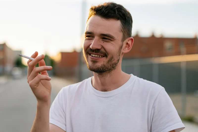 Young man smiling while smoking a cigarette outdoors on a street
