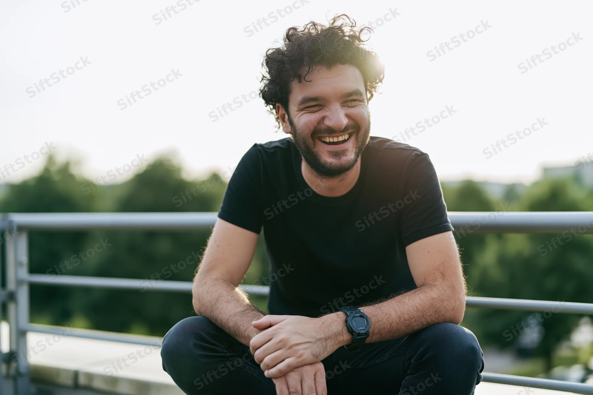 Young man laughing while sitting on an outdoor terrace with hands clasped and curly hair in sunlight