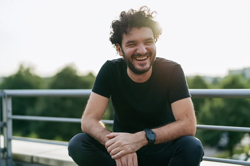 Young man laughing while sitting on an outdoor terrace with hands clasped and curly hair in sunlight