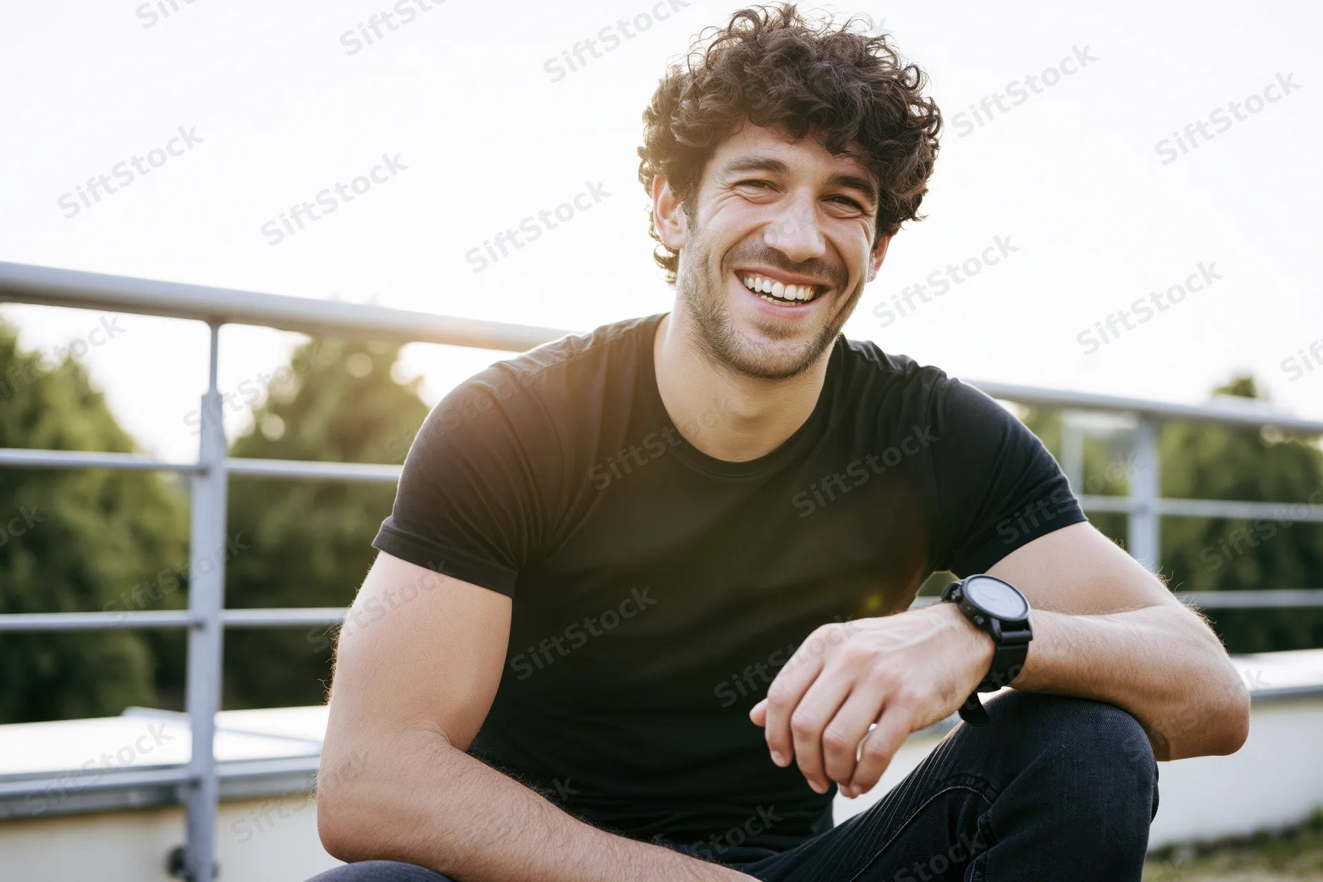 Young man with curly dark hair smiling while sitting on an outdoor terrace