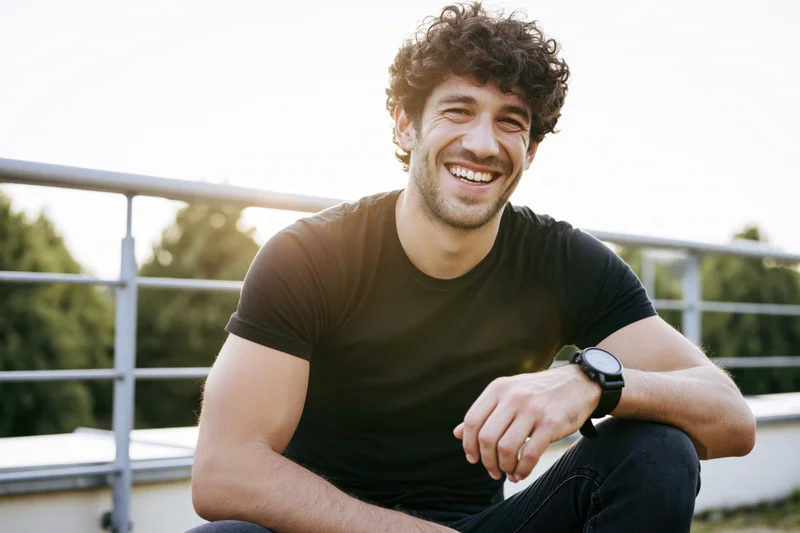 Young man with curly dark hair smiling while sitting on an outdoor terrace