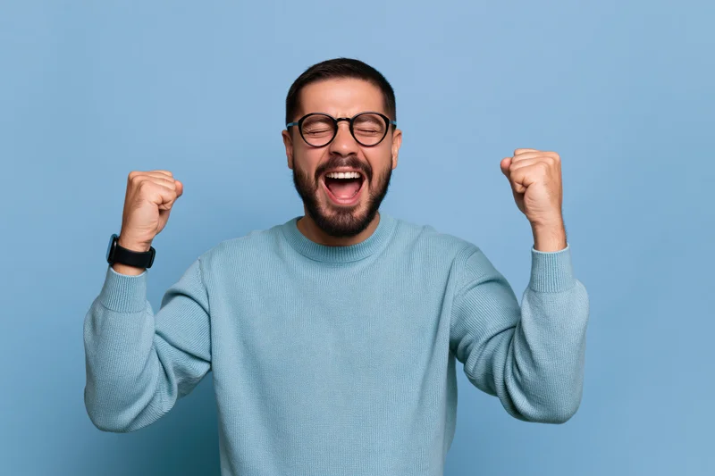 Young man with beard and glasses celebrating with raised fists against blue background