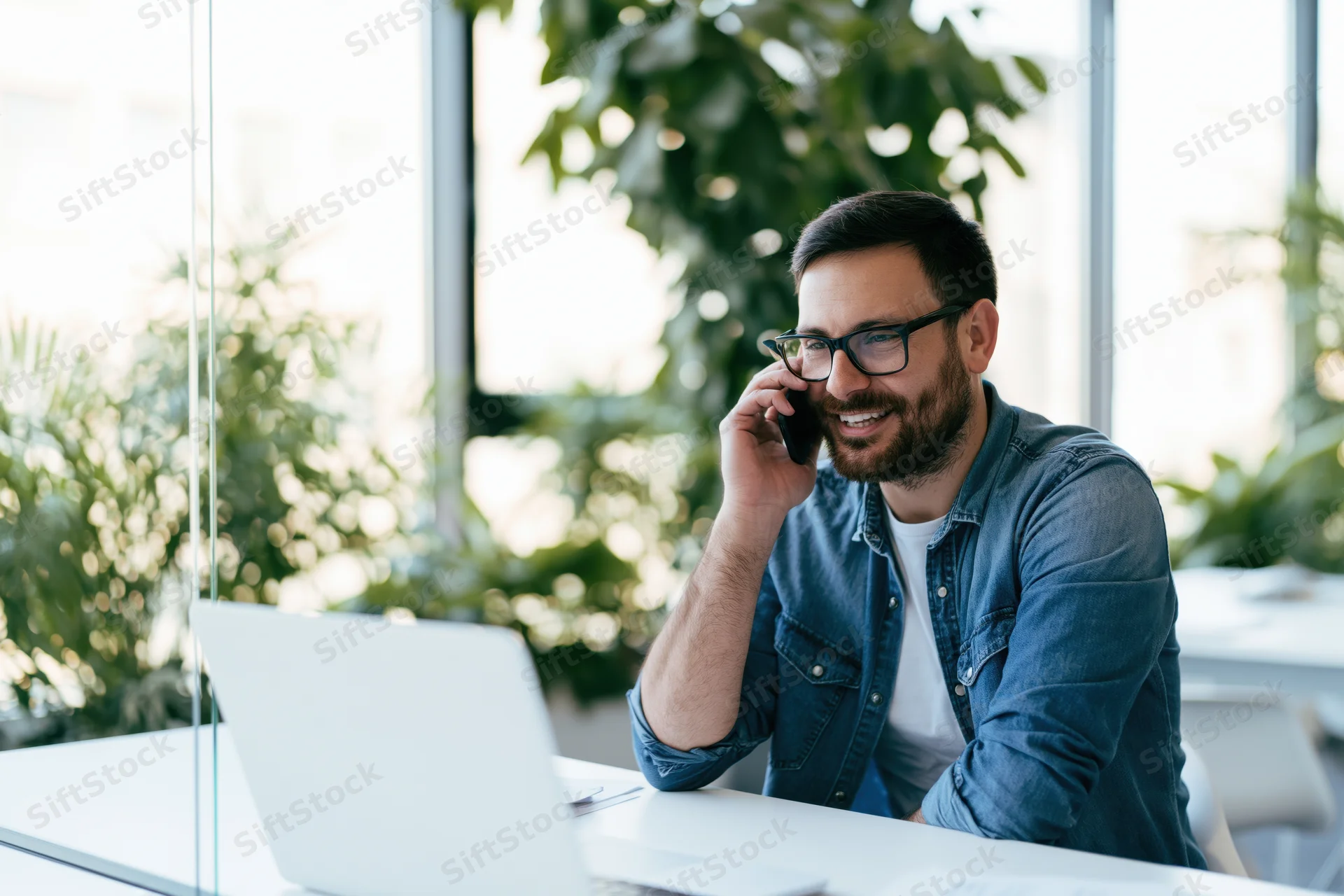Man smiling while talking on phone at desk with laptop in office