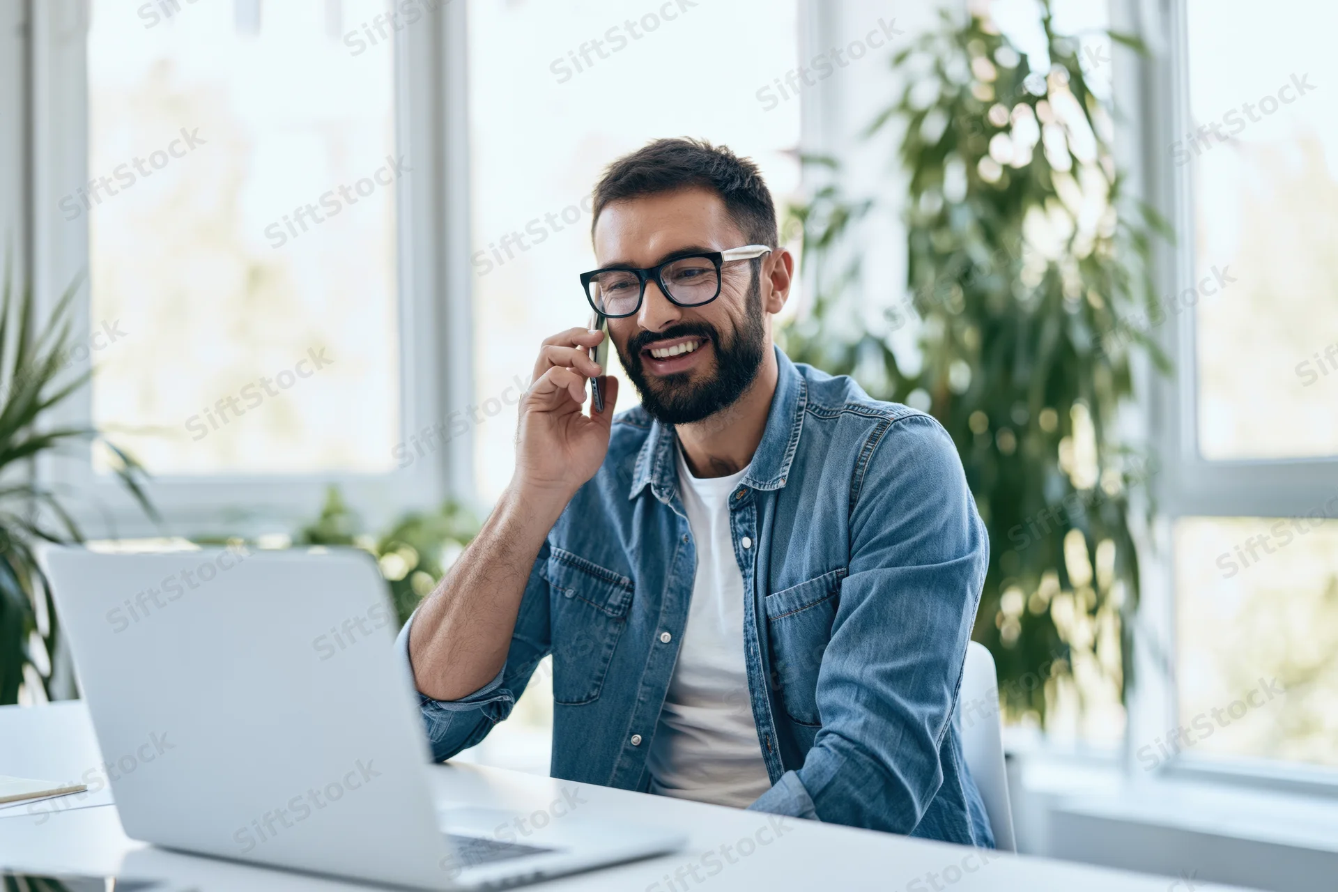 Man smiling while talking on smartphone at desk with laptop in bright office