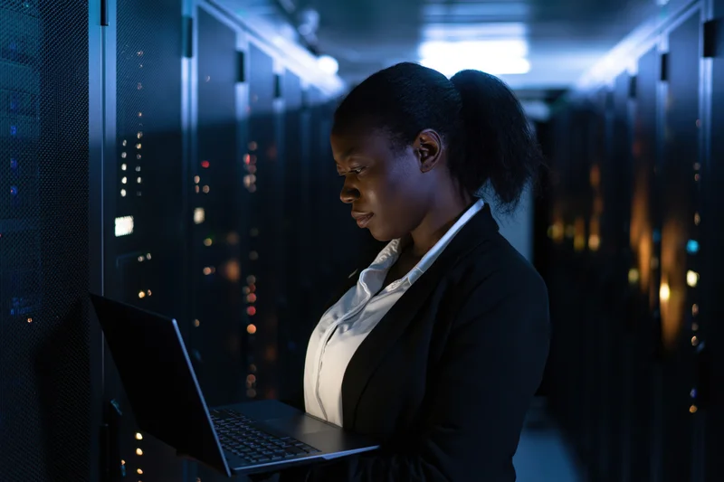 Woman technician using laptop in server room aisle
