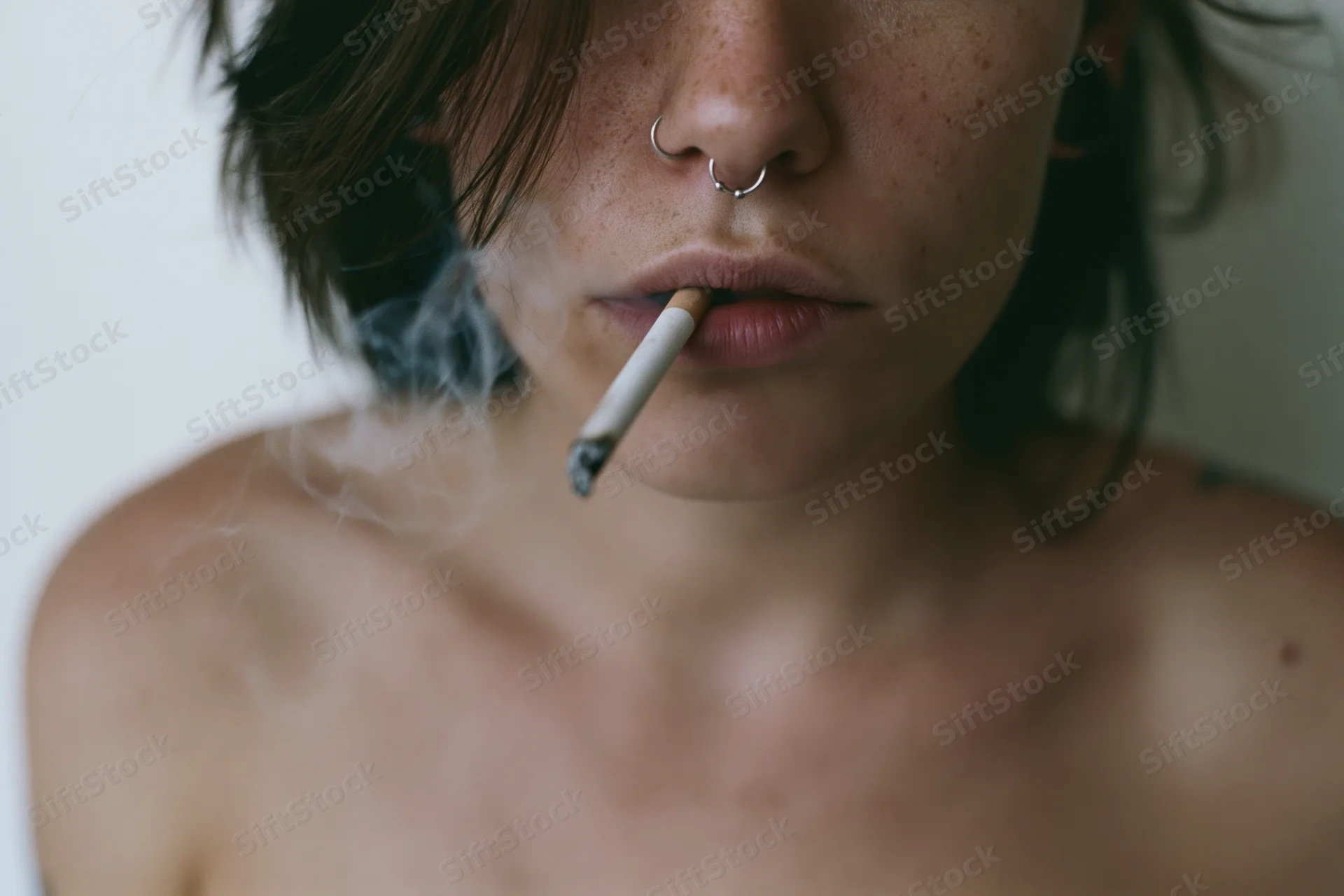Closeup of a young woman smoking a cigarette with smoke rising, bare shoulders visible against a plain white background