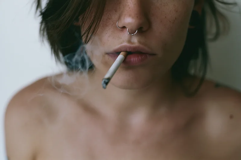 Closeup of a young woman smoking a cigarette with smoke rising, bare shoulders visible against a plain white background