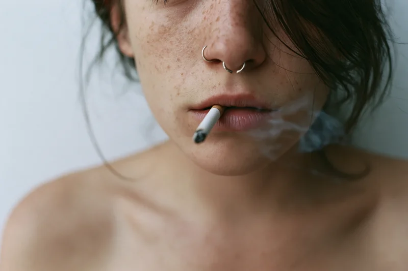 Close-up of a woman smoking a cigarette with smoke curling from her lips, bare shoulders visible against a neutral background