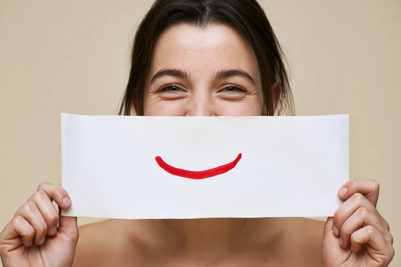 Young woman holding white paper with red smile drawn on it in front of her face