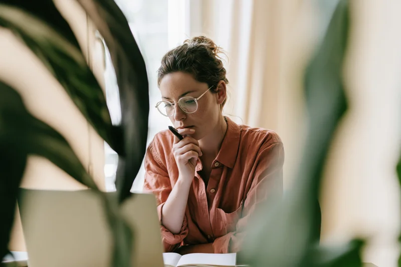 Woman thinking with pen to her chin while sitting at home desk surrounded by plants
